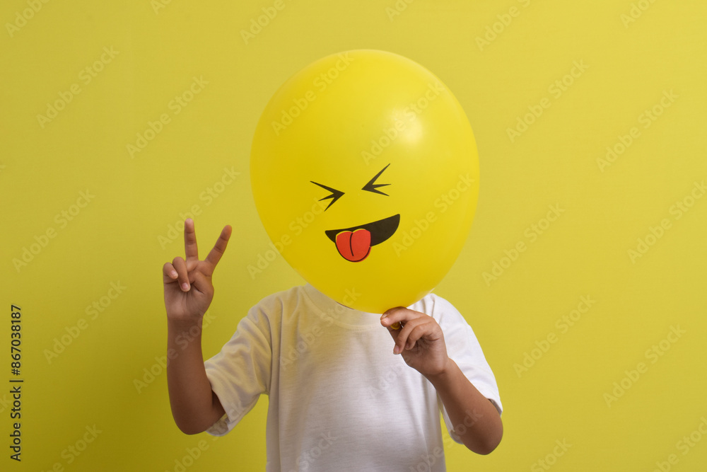 Tongue emoji. Cute Asian boy hiding his face behind balloon emoticon sticking out tongue isolated on yellow background. Teasing emoji with closed eyes Tongue emoji. Cute Asian boy hiding his face behind balloon emoticon sticking out tongue isolated on yellow background. Teasing emoji with closed eyes