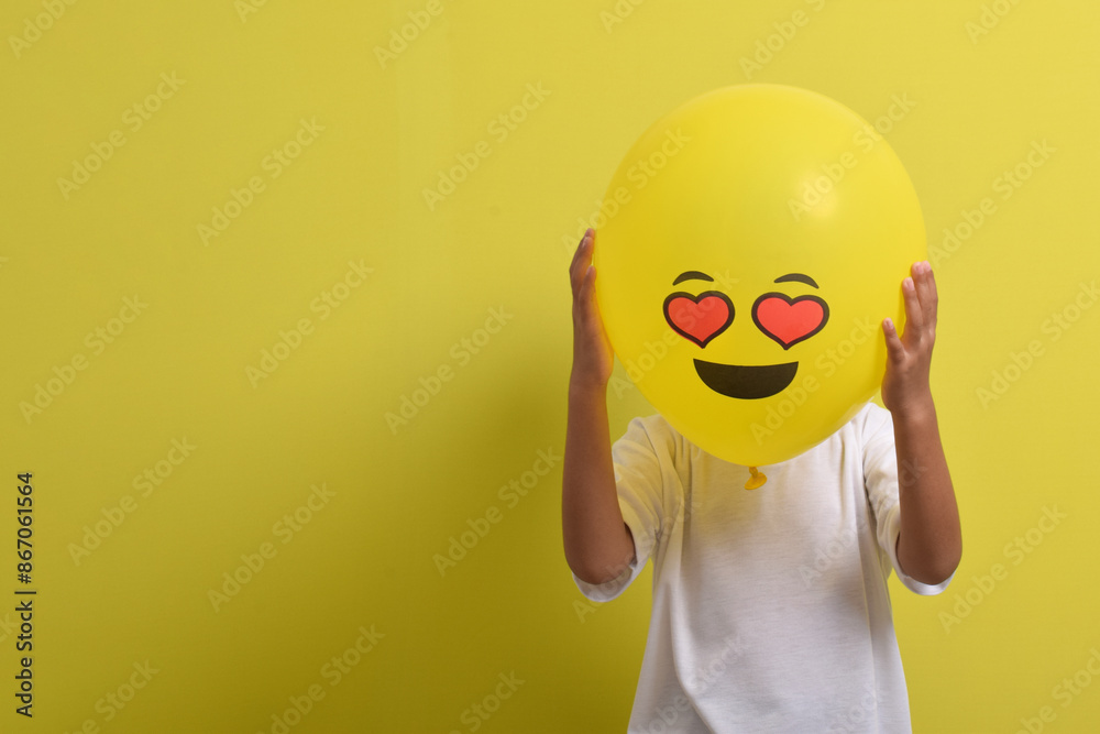 Emoticon in love. Cute Asian boy hides his face behind romantic love face emoticon balloons isolated on yellow background Emoticon in love. Cute Asian boy hides his face behind romantic love face emoticon balloons isolated on yellow background