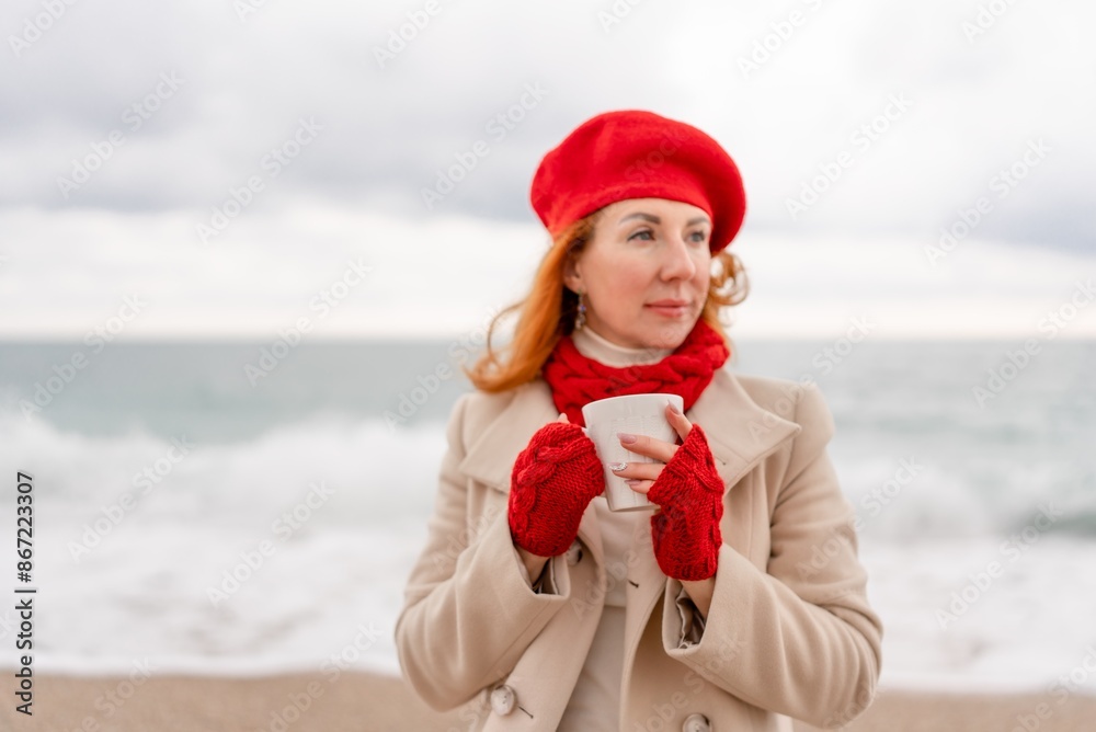 Obraz premium Woman beach in red beret scarf and mitts holding a cup of tea in his hands. Depicting beach relaxation and cozy attire. Walks by the sea
