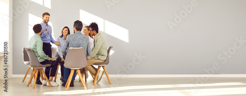 Business coach and team mentor meeting with group of people. Smiling man talking to happy young diverse men and women sitting in circle in modern office with gray copy space wall. Banner background
