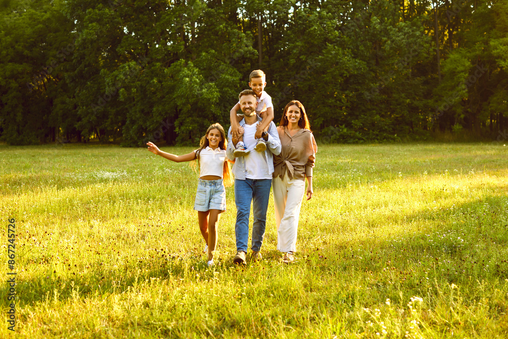 Fototapeta premium Portrait of happy smiling family with son and daughter standing in the summer park and looking cheerful at camera. Mother, father with two kids walking in nature enjoying sunny day together.