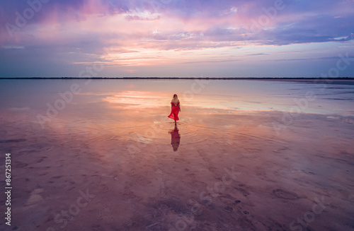 Aerial of a young woman in red dress walking in the water of a unique pink salt lake. Sunset at lake Bursol with beautiful reflections on calm water surface. Stunning scenery photo