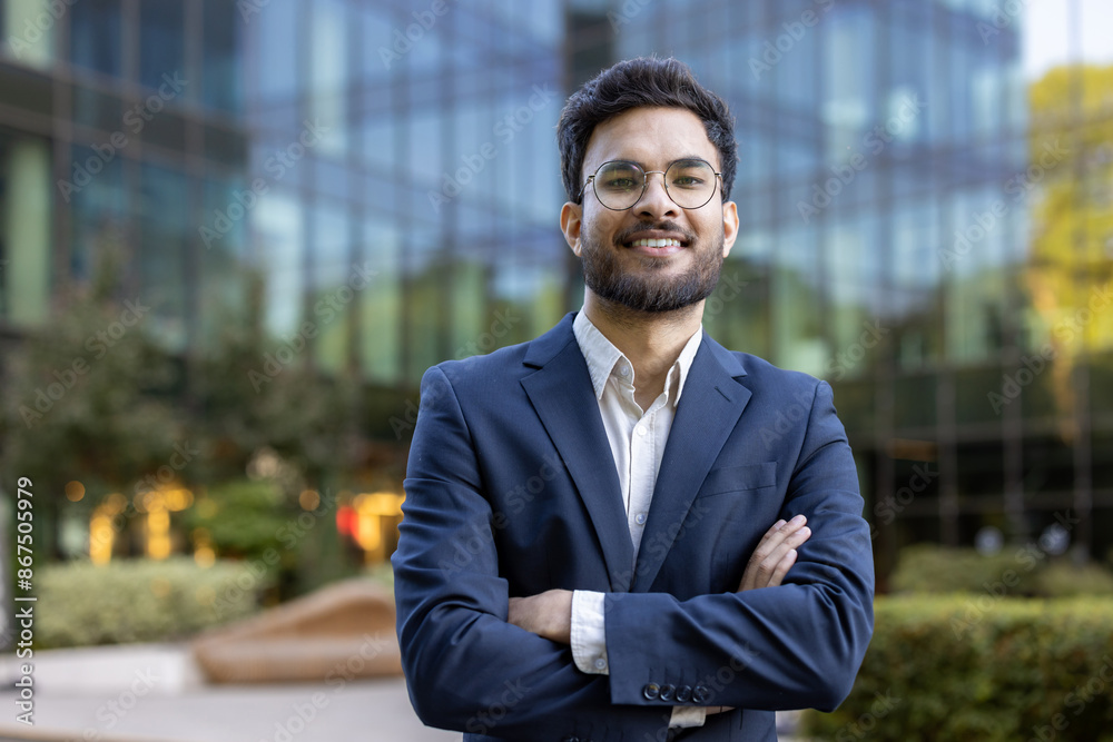 Confident young business professional standing outside modern office building with arms crossed and smiling. Business setting, professional appearance, outdoor background with glass windows Confident young business professional standing outside modern office building with arms crossed and smiling. Business setting, professional appearance, outdoor background with glass windows