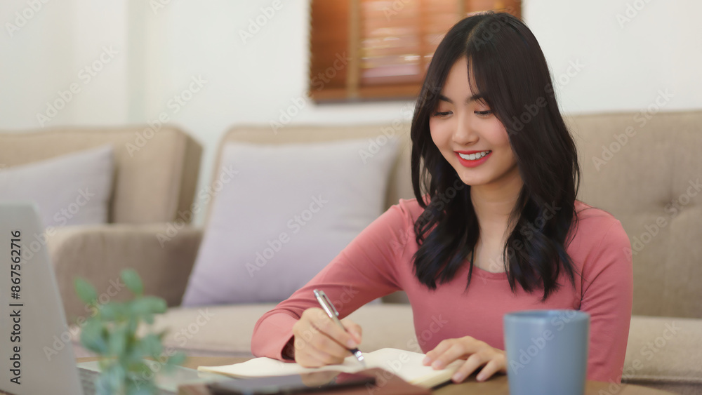 Young businesswoman reading data on laptop and writing notes in notebook while working at home Young businesswoman reading data on laptop and writing notes in notebook while working at home