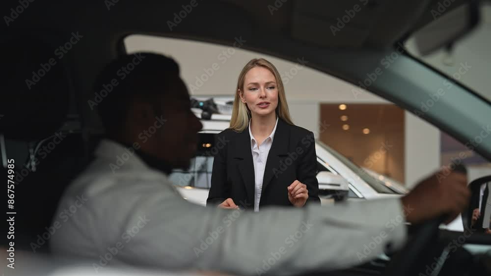 View from vehicle interior to positive female sales manager in business suit talking to black male client sitting in car at modern showroom. Happy African man sitting inside new car choose luxury auto View from vehicle interior to positive female sales manager in business suit talking to black male client sitting in car at modern showroom. Happy African man sitting inside new car choose luxury auto