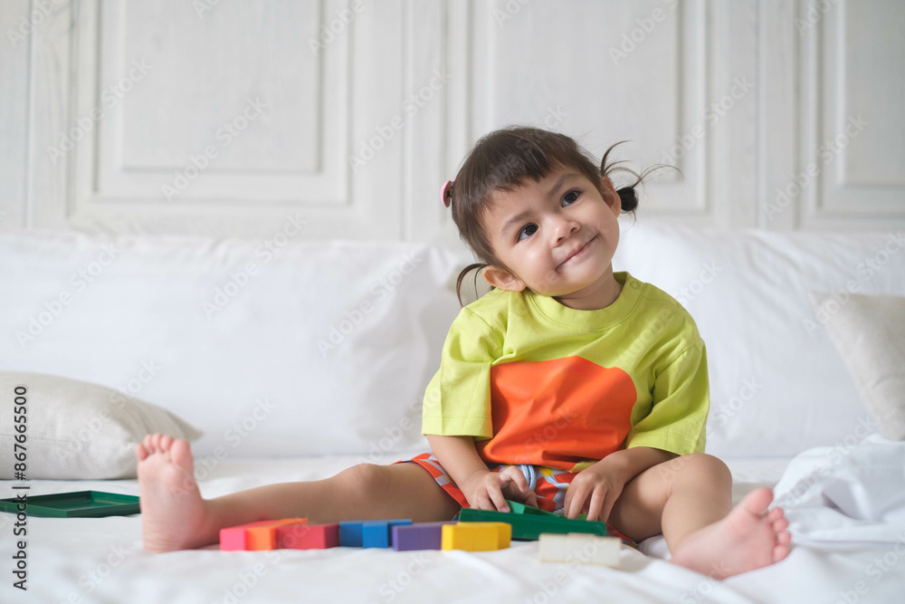 Fototapeta premium Adorable child sitting on a bed with colorful blocks, smiling and enjoying playtime in a bright and cozy room.