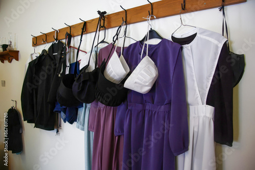 Amish clothing on wooden rack: plain, modest garments. Purple dresses, black coats, white bonnets. Represents Amish culture, traditional religious attire, simple living, and faith-based dress codes. photo