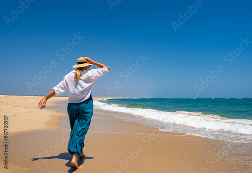 Summer vacation. Beautiful young woman in white blouse and baggy pants fluttering in the wind walking on a sandy beach by the sea with big waves on a beautiful sunny day