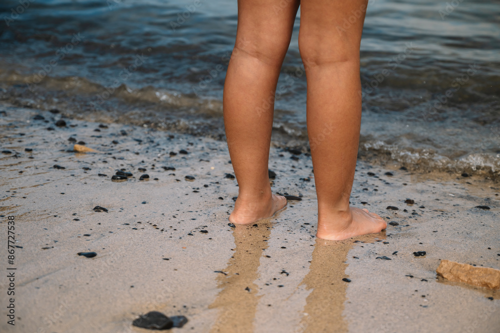 A person standing in the sand with their feet in the water. The water is dark and murky, and the sand is littered with rocks A person standing in the sand with their feet in the water. The water is dark and murky, and the sand is littered with rocks