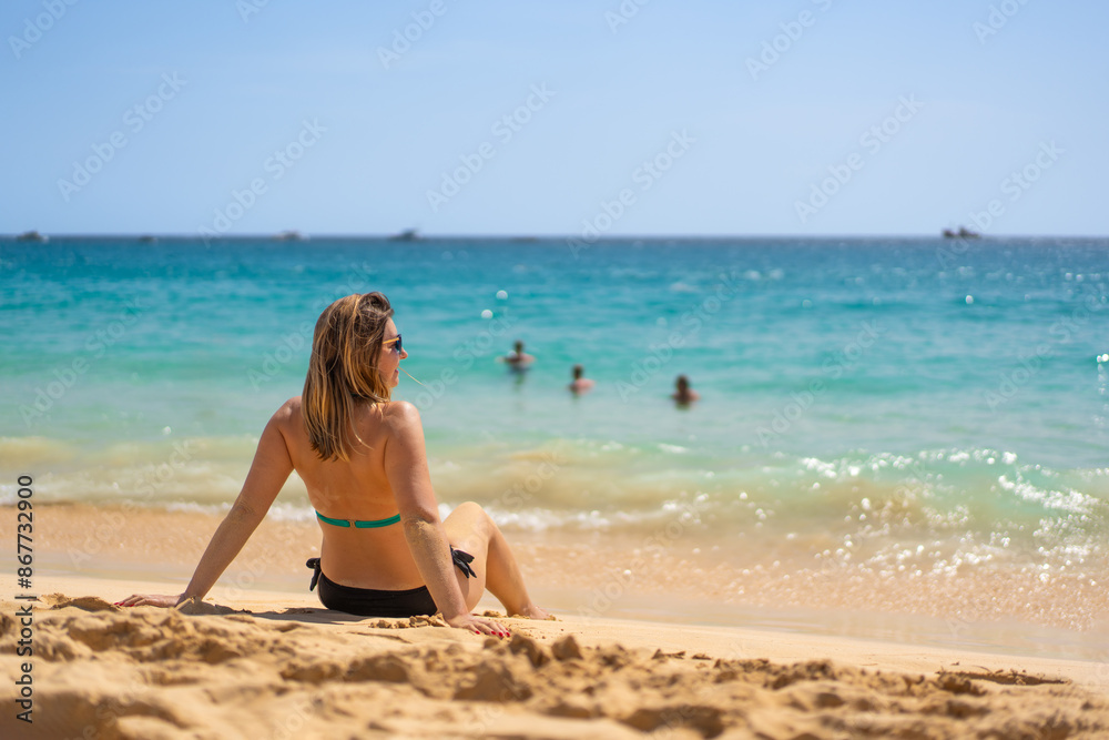 Beach vacation. Happy young woman in bikini sitting on sandy beach by the sea on beautiful sunny day. Positive emotion Beach vacation. Happy young woman in bikini sitting on sandy beach by the sea on beautiful sunny day. Positive emotion