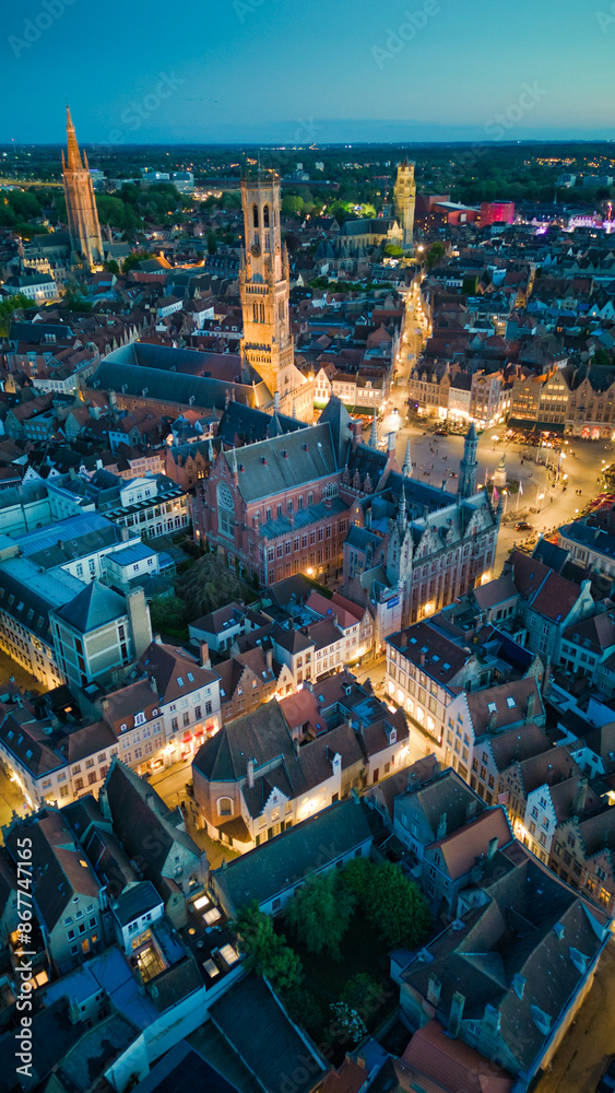 Naklejka premium Enchanting aerial view of Bruges' historic old town captured by a drone at night! The illuminated medieval architecture, winding canals, and charming cobblestone streets create a magical and captivati