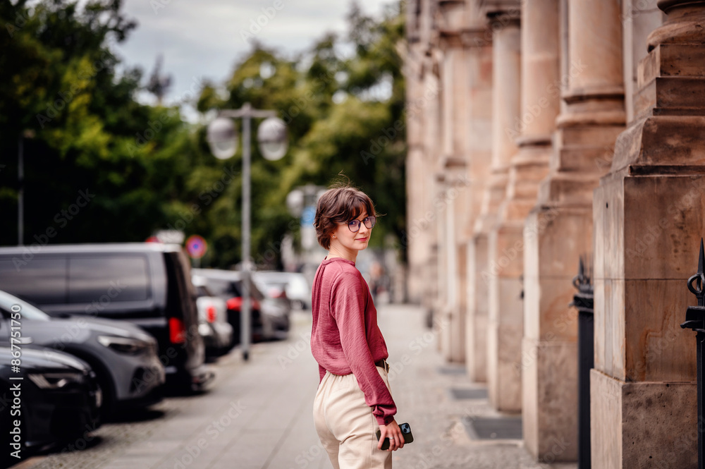 Woman in casual attire walking on a city sidewalk holding her phone, surrounded by parked cars and historic buildings.