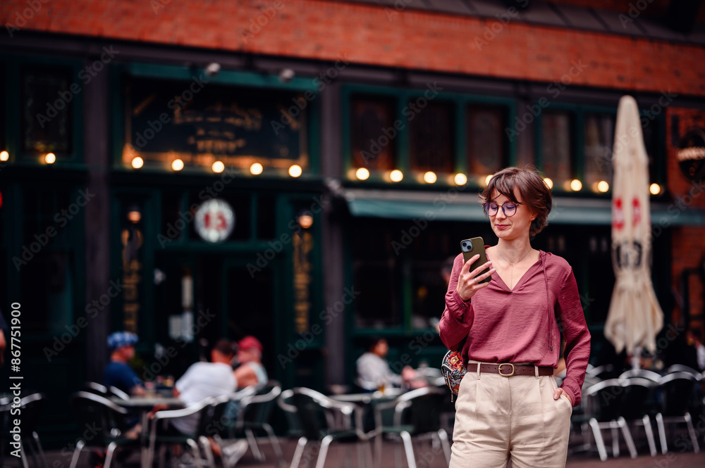 Obraz premium Woman in casual attire checking her phone while standing outside a busy cafe with people seated at tables.