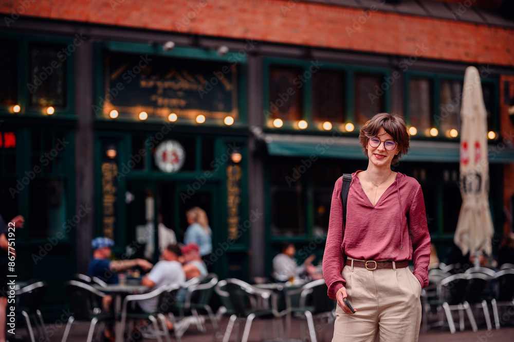 Fototapeta premium Woman in casual attire smiling and holding her phone while standing outside a cafe with people sitting at tables.