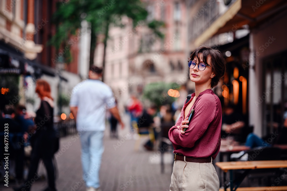 Woman in casual attire and glasses, smiling confidently with her phone and backpack, standing in a bustling city street lined with cafes and people. Woman in casual attire and glasses, smiling confidently with her phone and backpack, standing in a bustling city street lined with cafes and people.