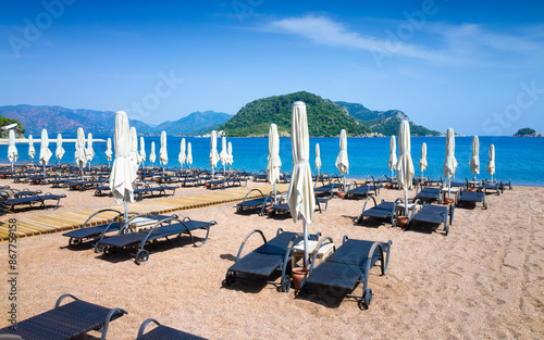 Фотография Icmeler Beach in Marmaris, Turkey with rows of folded white parasols and sun loungers on sandy shore, ready for tourists to enjoy calm blue waters