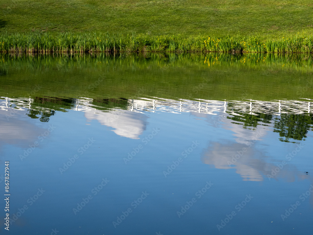 Fototapeta premium A Virginia neighborhood pond mirrors the sky, clouds, and a white fence in its tranquil waters.