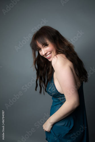 Studio portrait of a beautiful caucasian woman wearing a royal blue nightgown. She has long wavy brown hair. She is laughing and looking over her shoulder. The background is grey.