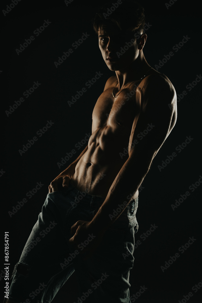 Muscular man posing shirtless in dark moody lighting, showcasing his defined abs and physique, wearing ripped jeans in a dramatic, mysterious studio setting. Muscular man posing shirtless in dark moody lighting, showcasing his defined abs and physique, wearing ripped jeans in a dramatic, mysterious studio setting.