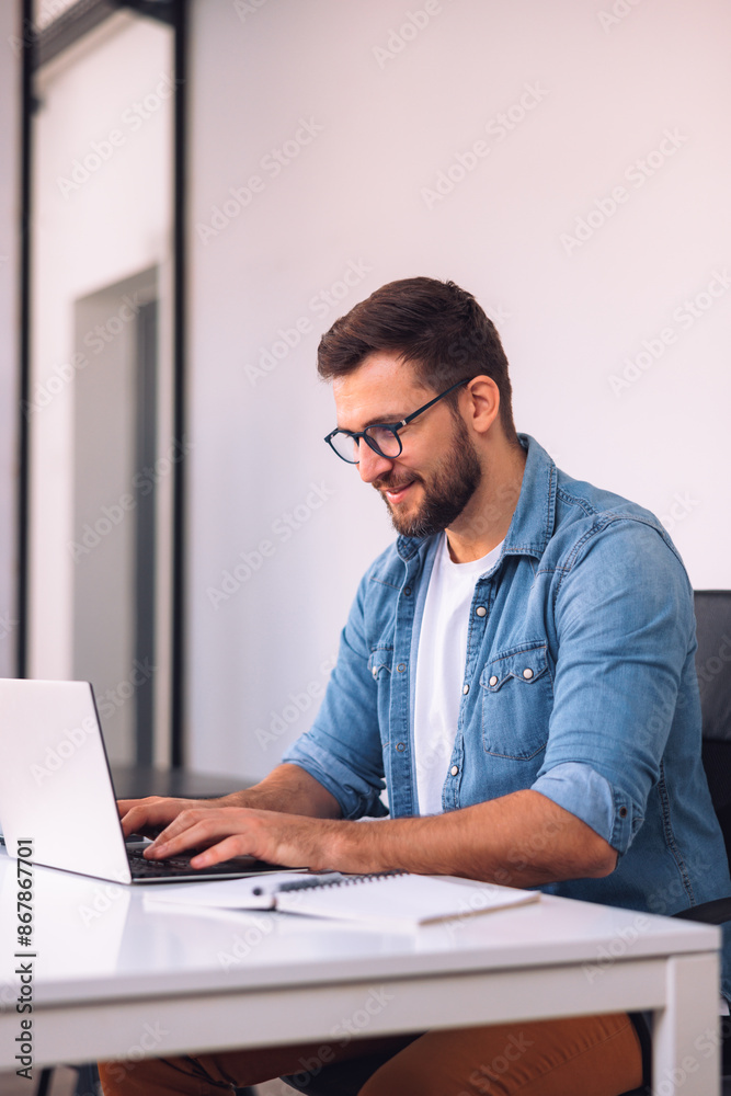 A smiling man with glasses is using a laptop in the office while sitting at his desk. A smiling man with glasses is using a laptop in the office while sitting at his desk.