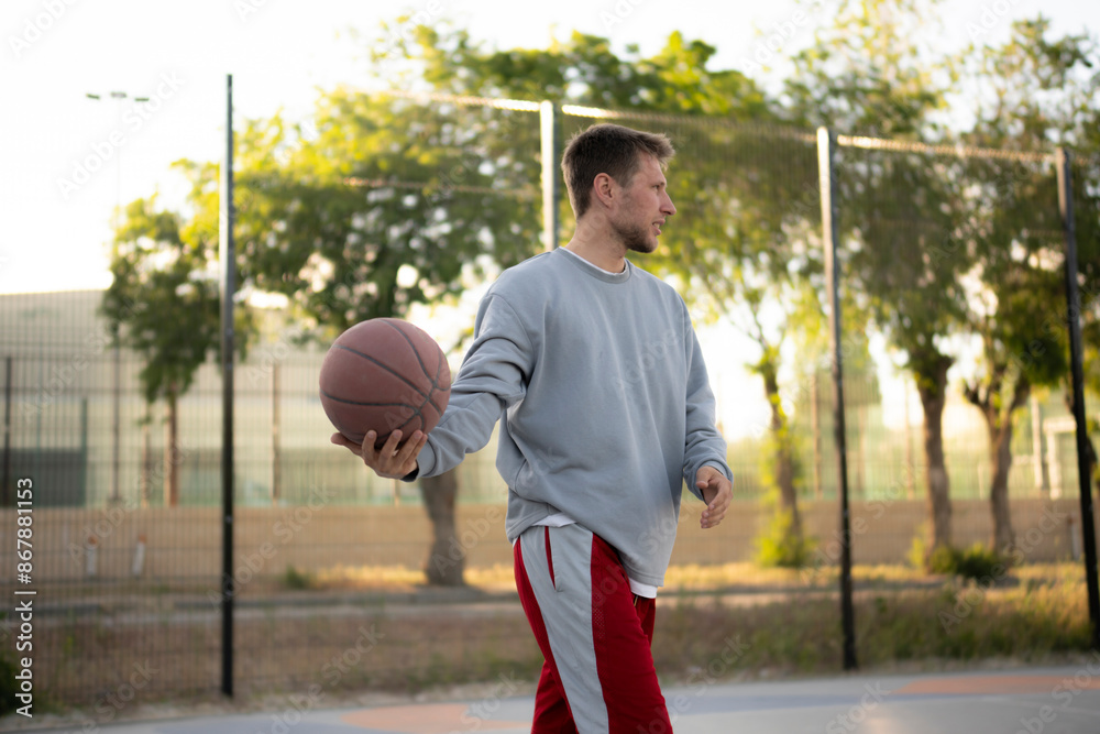 young man stands on a basketball court, holding a basketball in his hands. The sun shines brightly above him, casting a warm glow on the court and creating long shadows from the trees young man stands on a basketball court, holding a basketball in his hands. The sun shines brightly above him, casting a warm glow on the court and creating long shadows from the trees