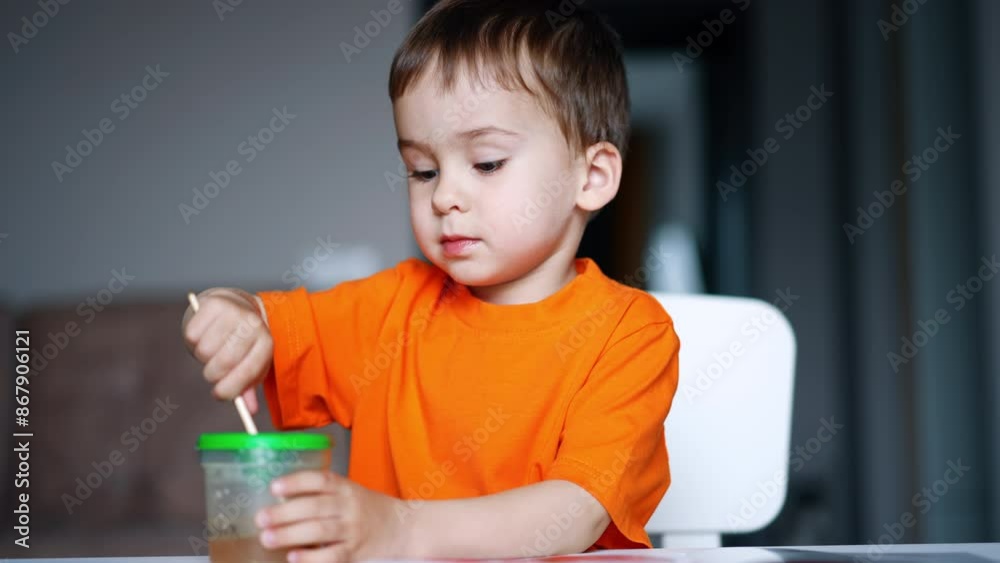 Little boy carefully washes his paint brush in the cup. Mom's hand points at the paper and kid starts to paint. Blurred backdrop. Little boy carefully washes his paint brush in the cup. Mom's hand points at the paper and kid starts to paint. Blurred backdrop.