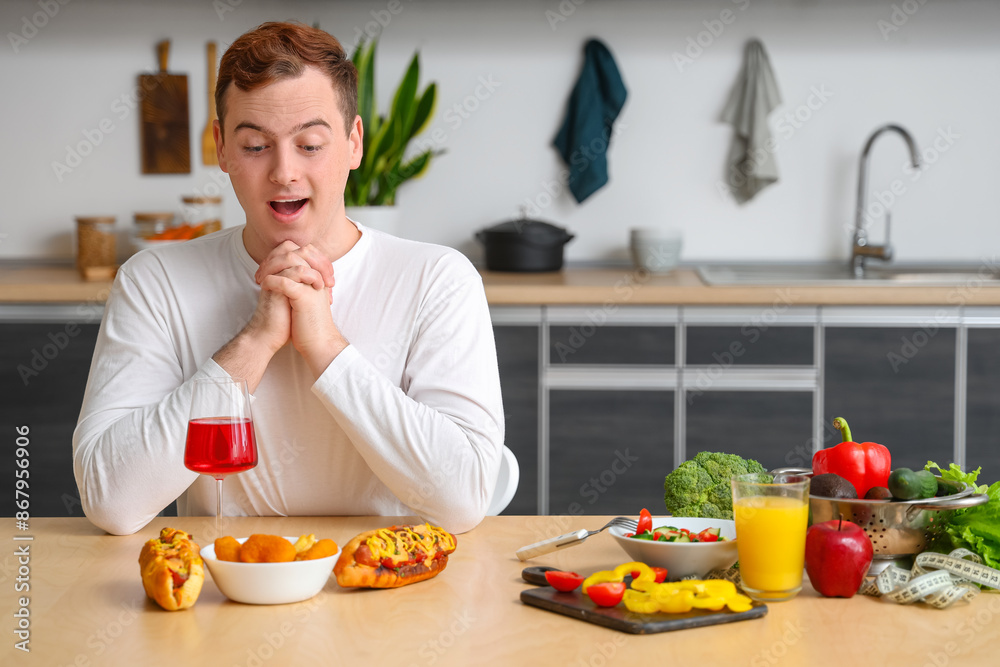 Young shocked overweight man choosing fast food over healthy food at table in kitchen. Weight gain concept Young shocked overweight man choosing fast food over healthy food at table in kitchen. Weight gain concept