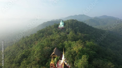 Aerial view of a temple nestled in the forest with a large green Buddha statue on a mountain peak. Tranquil spiritual scene in Thailand.
