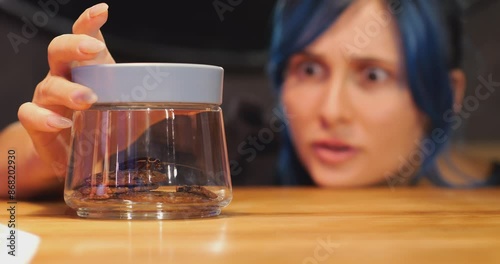 Woman found cockroach in closed glass jar with cookies, look to pest with disgust. Selective focus on container with few snacks on bottom and roach sitting on them, blurred face on background photo