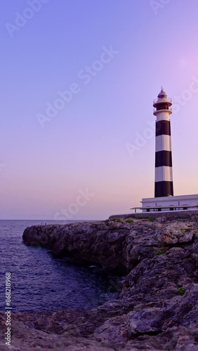 Vertical video of the iconic Artrutx Lighthouse situated on the rocky coast at sunset. The tranquil sea and pastel sky create a serene and picturesque scene, perfect for travel to the island of photo