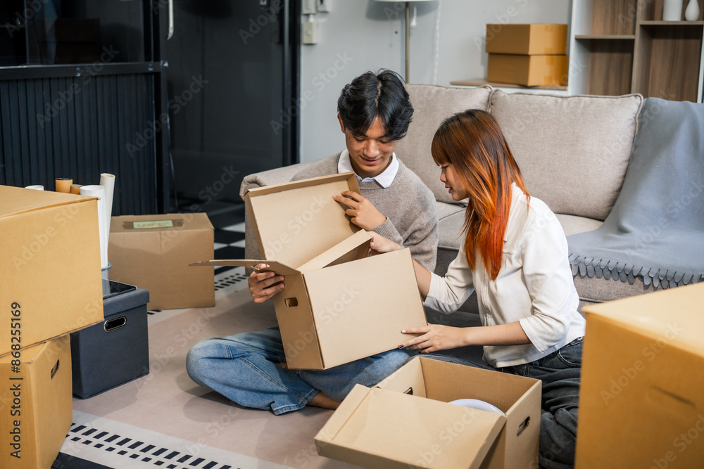 Indoors, a young couple happily packs cardboard boxes, preparing for a move. They share smiles and excitement, embracing new beginnings together in their new home, filled with love and anticipation