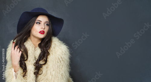 Stylish fashion model woman with long dark wavy hairstyle and red lips make-up wearing fashionable hat beauty studio portrait © millaf