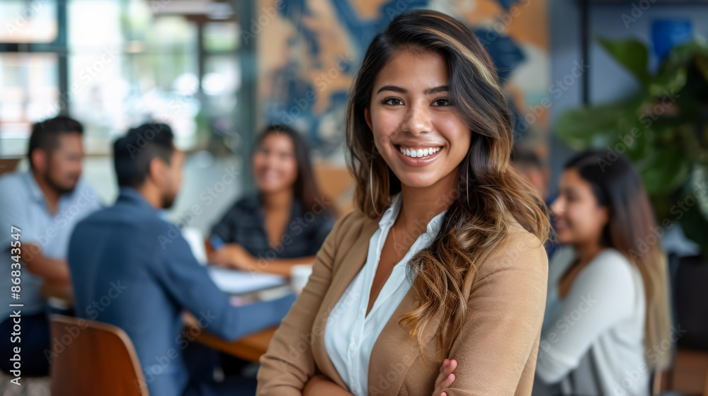 A bright photo of a young Latina woman with long dark hair, standing and smiling confidently with her arms crossed, while colleagues discuss in the background. A bright photo of a young Latina woman with long dark hair, standing and smiling confidently with her arms crossed, while colleagues discuss in the background.