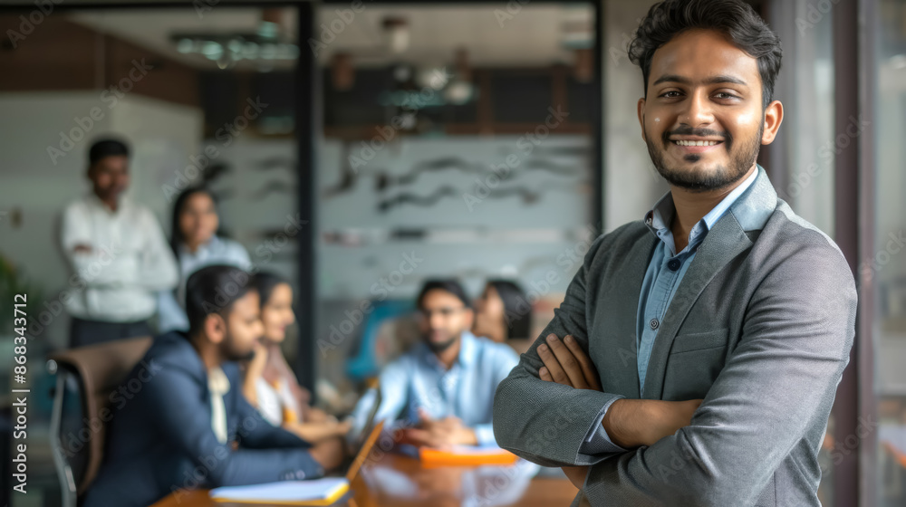 A photo of a young Indian man with short black hair, wearing a grey blazer, standing with arms crossed in a modern office, while his colleagues are in a meeting in the background. A photo of a young Indian man with short black hair, wearing a grey blazer, standing with arms crossed in a modern office, while his colleagues are in a meeting in the background.