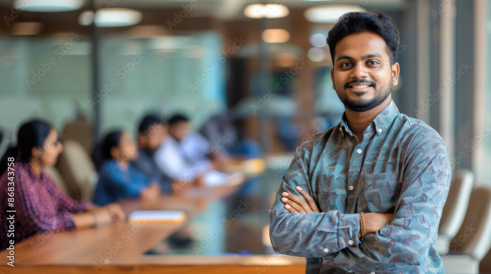 A photo of a young Indian man with short black hair, smiling with arms crossed in a modern conference room, while colleagues discuss at a table in the background. A photo of a young Indian man with short black hair, smiling with arms crossed in a modern conference room, while colleagues discuss at a table in the background.