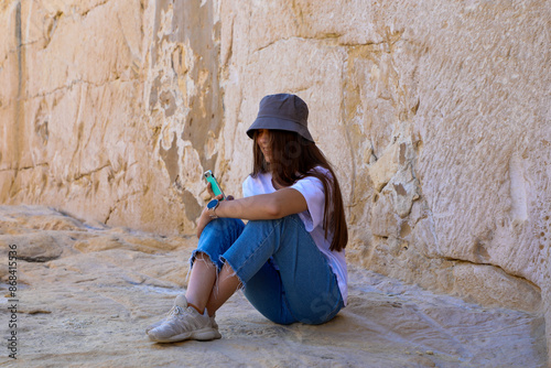 A teenage girl in blue jeans with her hair down, with a mobile phone in her hand and a hat on her head, sits on the rocks and smiles. © Svitlana
