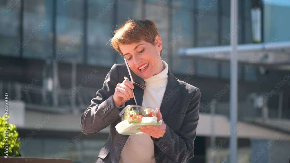A woman in a business suit is eating a salad while talking on her cell phone. Concept of busyness and multitasking, as the woman is trying to eat and talk at the same time A woman in a business suit is eating a salad while talking on her cell phone. Concept of busyness and multitasking, as the woman is trying to eat and talk at the same time