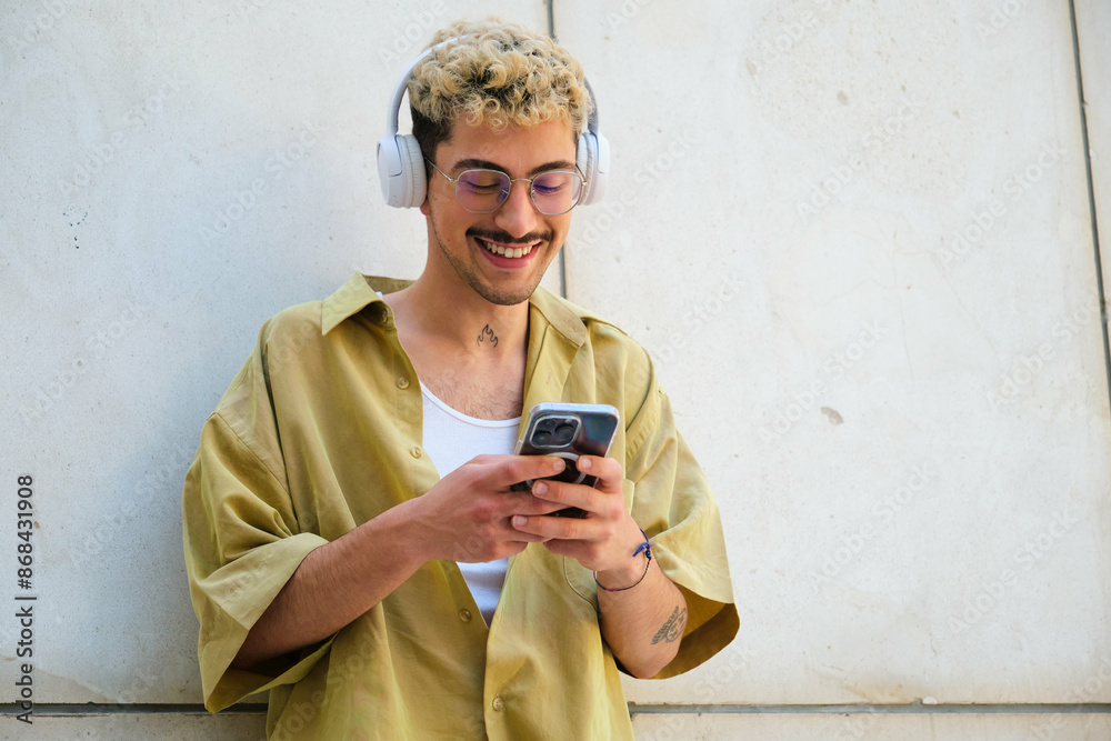 A man with a beard and glasses is smiling while holding a cell phone. He is wearing headphones and he is listening to music