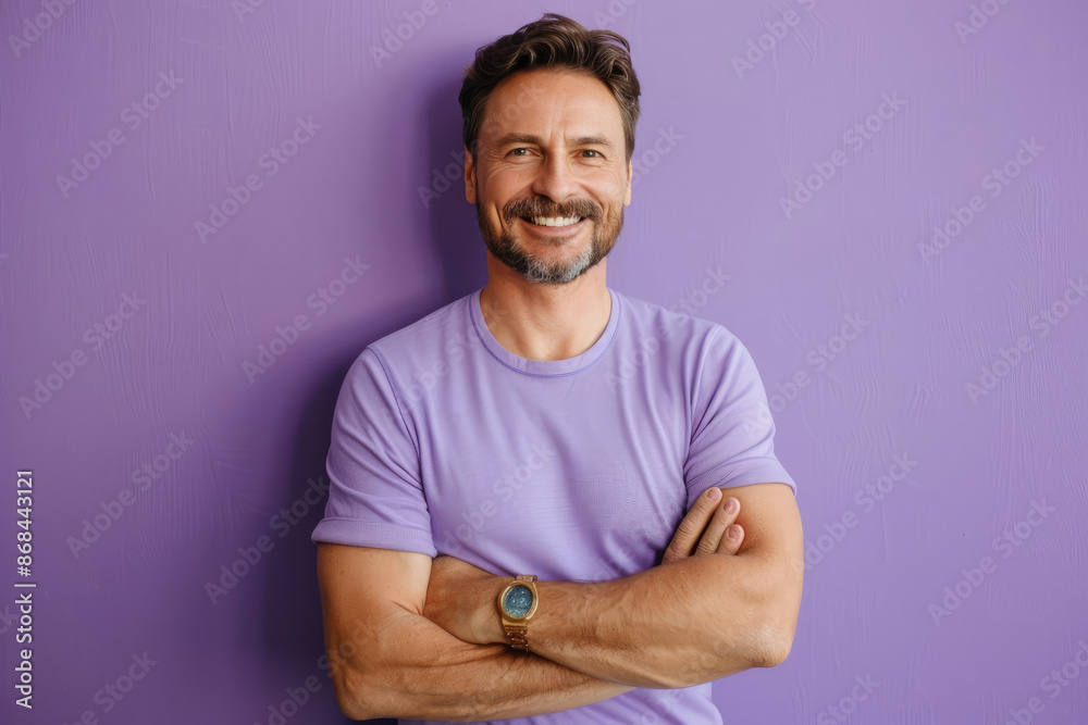 A confident man with a charming smile stands with arms crossed, sporting a watch, against a solid purple background