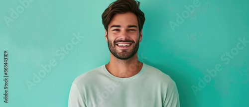 A young man with stubble and a trendy haircut, smiling earnestly, against a solid teal colored wall
