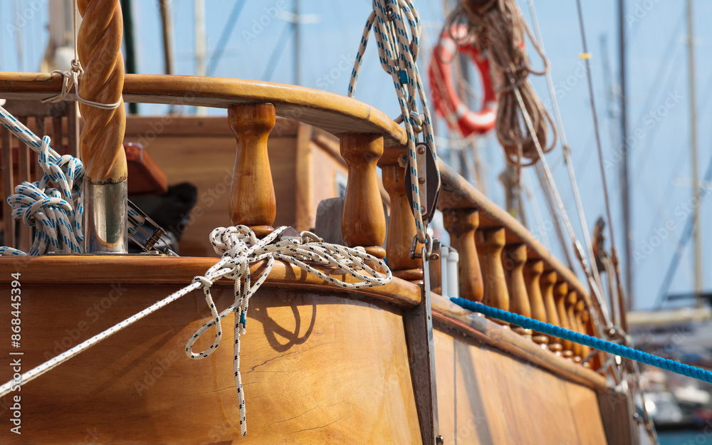 Obraz premium Intricate details of sailing boat’s deck, featuring neatly coiled ropes and polished wood, with other vessels’ masts in soft-focus background, evoking spirit of maritime tradition
