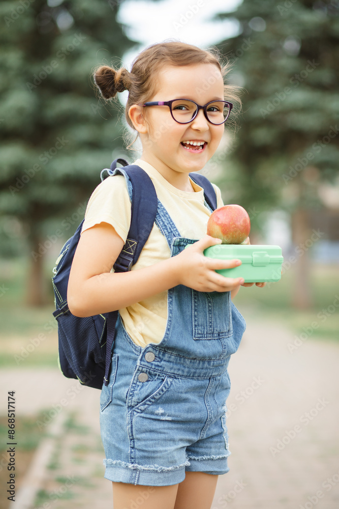 Cute little schoolgirl with backpack holding in a hands lunch box and apple. Near by school outdoor. Food for kids. Cute little schoolgirl with backpack holding in a hands lunch box and apple. Near by school outdoor. Food for kids.