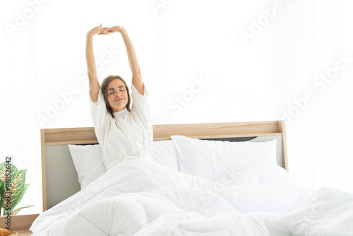 Woman stretching in bed after wake up. she sitting near the big white window while stretching on bed after waking up with sunrise at morning