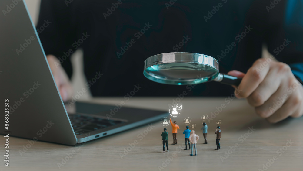 A close-up of a person's hand holding a magnifying glass over a group of miniature figures, individual. targeted search, recruitment, and selection processes in a professional or business context.