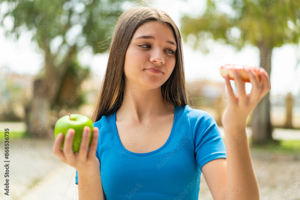 Teenager girl at outdoors holding apple and donut and thinking Teenager girl at outdoors holding apple and donut and thinking