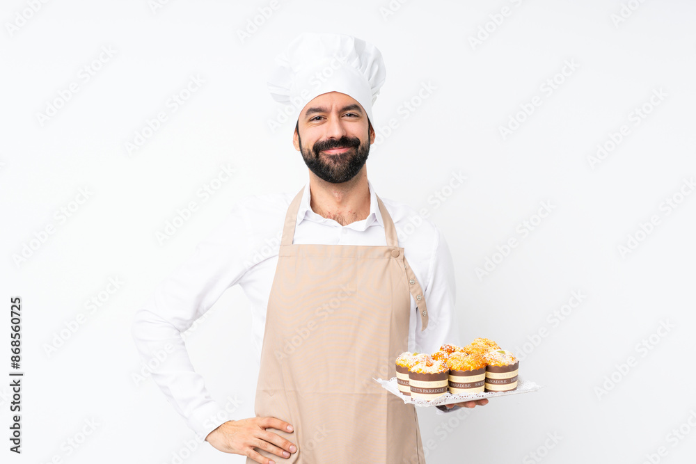 Young man holding muffin cake over isolated white background posing with arms at hip and smiling Young man holding muffin cake over isolated white background posing with arms at hip and smiling