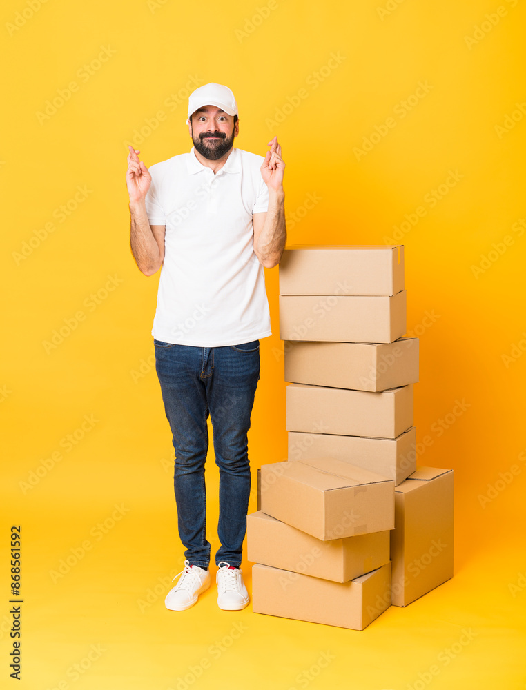 Full-length shot of delivery man among boxes over isolated yellow background with fingers crossing and wishing the best Full-length shot of delivery man among boxes over isolated yellow background with fingers crossing and wishing the best