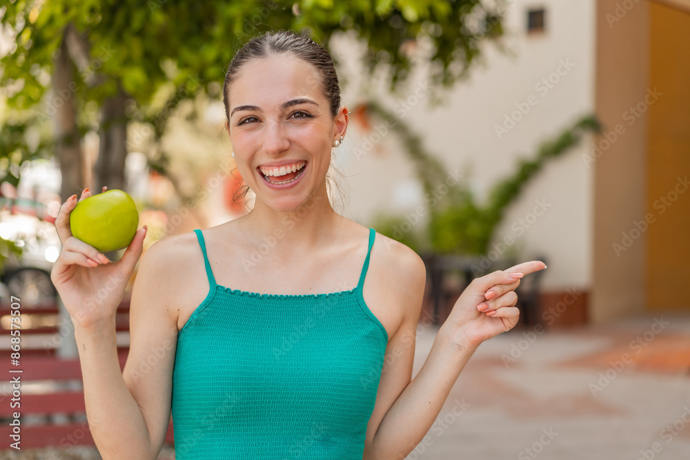 Young pretty woman with an apple at outdoors surprised and pointing finger to the side Young pretty woman with an apple at outdoors surprised and pointing finger to the side