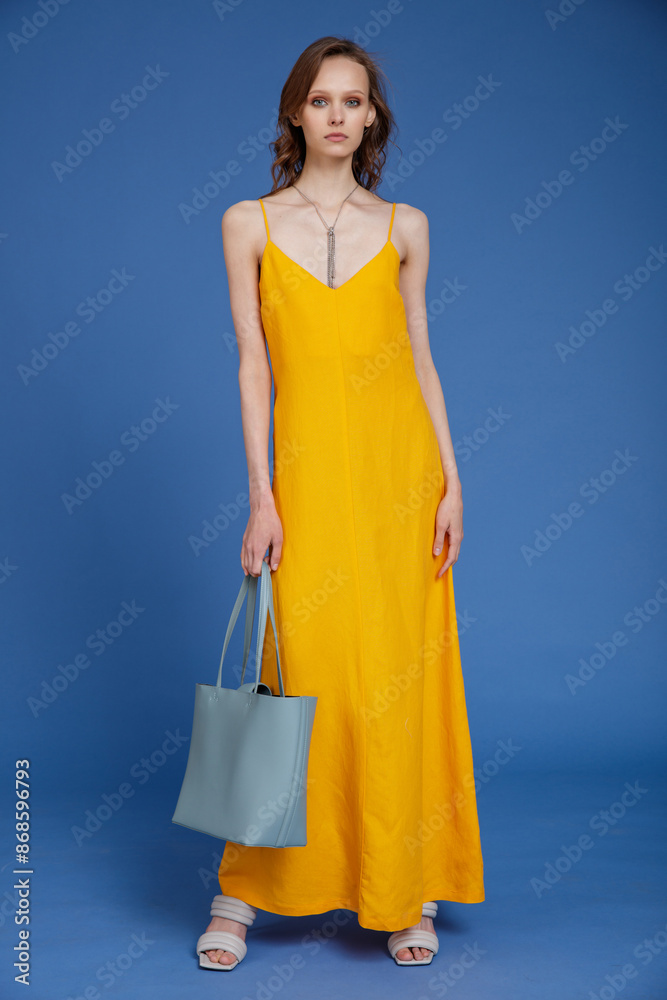 High fashion photo of a beautiful skinny young woman in pretty long yellow dress, handbag posing on blue background. Slim figure. High fashion photo of a beautiful skinny young woman in pretty long yellow dress, handbag posing on blue background. Slim figure.