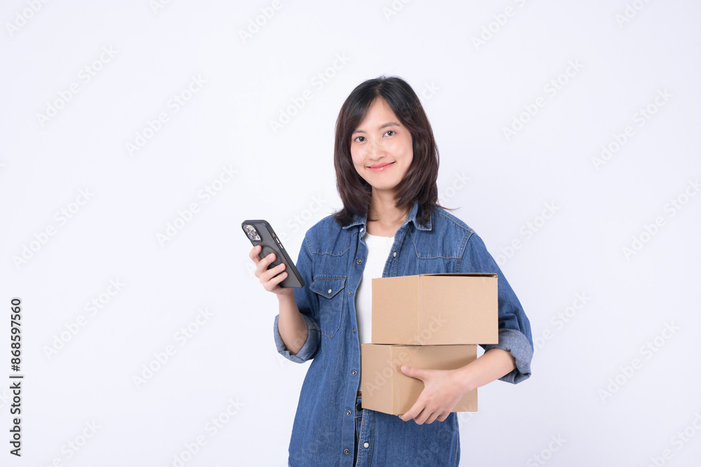 asian woman wearing denim jean holding parcel box while using mobile phone isolated on white studio background, Delivery courier and shipping service concept. asian woman wearing denim jean holding parcel box while using mobile phone isolated on white studio background, Delivery courier and shipping service concept.
